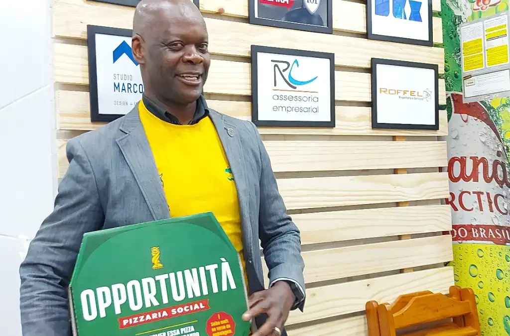 Homem sorrindo segura a caixa verde da Opportunità Pizzaria Social, com a mensagem de que ao comer a pizza você colabora com projetos sociais