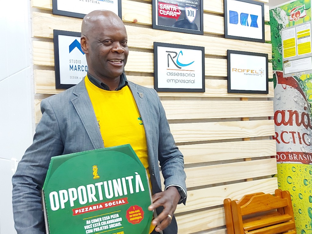 Homem sorrindo segura a caixa verde da Opportunità Pizzaria Social, com a mensagem de que ao comer a pizza você colabora com projetos sociais
